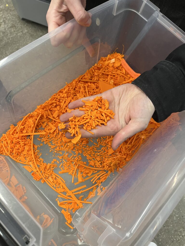 A clear bucket with orange shreds of PLA 3D printer filament.