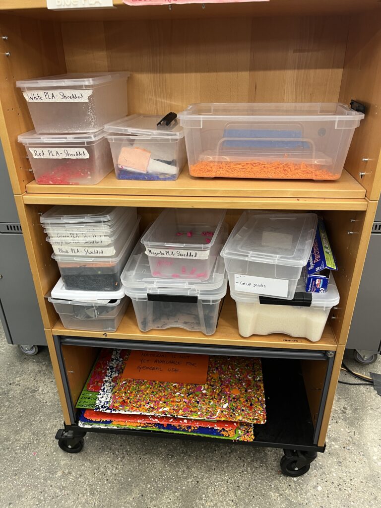 A shelf holds clear plastic bins of shredded 3D printer filament sorted by color.