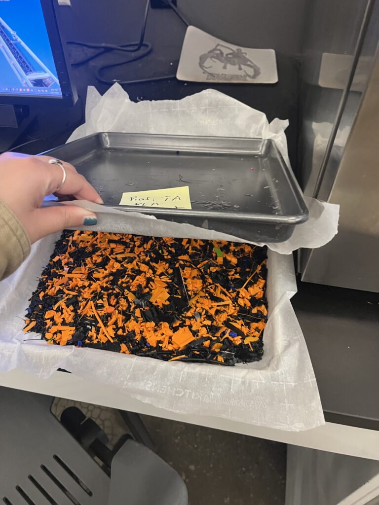 A sheet of melted together shreds of orange and black 3D printer filament is cooling between two baking trays.