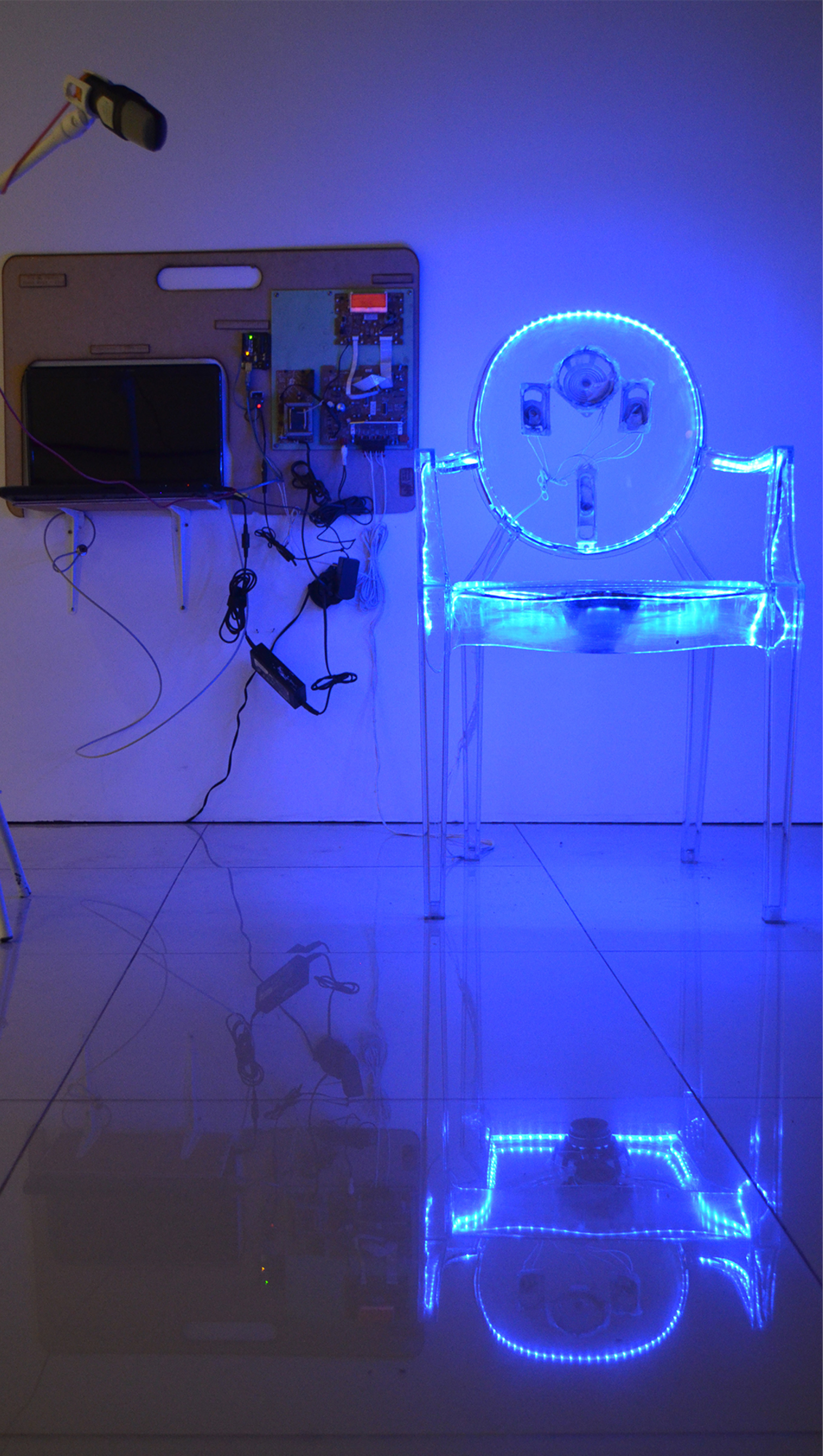 Photo of 'Inside Voice' (2018) displaying a microphone next to a transparent acrylic chair lit up in blue. In the back, there is a panel on a wall holding a computer and a set of wires and electronics. (Photo: Carina Macedo)