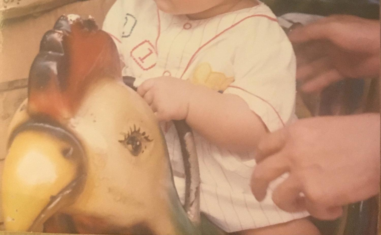 A toddler riding a mechanical chicken, with two adult hands nearby.