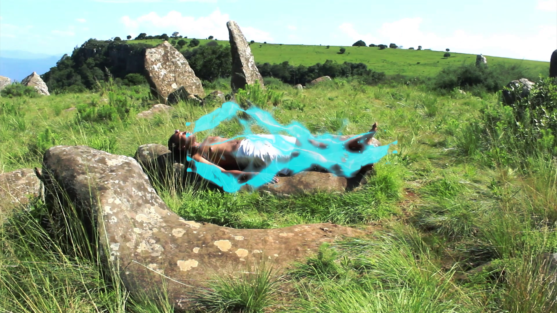 A person, covered in a bluish glow, lying in the grass, surrounded by rocks.