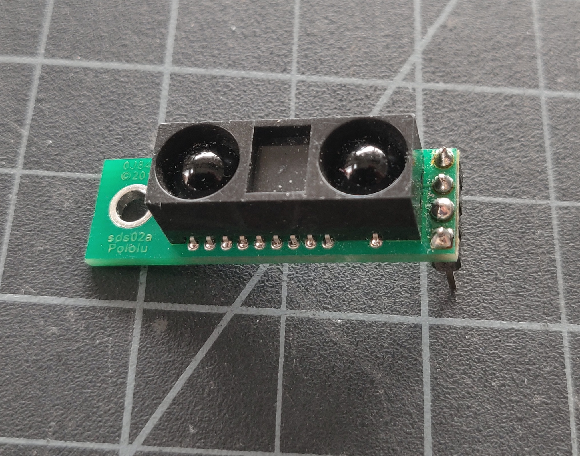 Photo of an infrared distance sensor. It has two lenses on top, one for the light and one for the sensor, and three pins on the side, to plug into a solderless breadboard. 