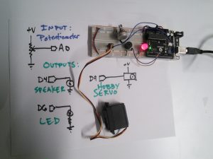 Complete photo of circuit with speaker, LED, and servo on breadboard with whiteboard drawing of schematic of presented schematic.