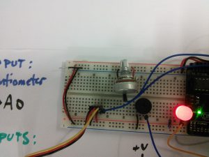Photo closeup of circuit with speaker, LED, and servo on breadboard with whiteboard drawing of schematic of presented schematic.