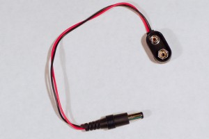 9V battery snap. This flat component with rounded ends has two round metal mounts on one side. One is slightly bigger than the other, about half an inch (3-2cm) across. They are designed to snap onto a 9-volt battery. There is a pair of wires coming out one end that has a DC power plug attached to it. This can plug into a DC power jack.