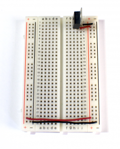 A solderless breadboard with a 7805 5-Volt voltage regulator mounted on it. There are several rows of holes for components. The holes on the breadboard are separated by 0.1-inch spaces, and are organized in many short rows in the center, and in two long rows down each side of the board. The short horizontal rows in the middle are separated by a center divider. The side rows are usually marked red, for voltage, and blue or black, for ground. On this breadboard, the side rows on the left side are connected to their counterparts on the right side with wires. The voltage regulator is mounted in the top three short rows on the right side of the board. The regulator faces to the left. The row holding the regulator's center pin is connected to the blue or black side row on the right side. The row holding the regulator's bottom pin is connected to the red side row on the right side.