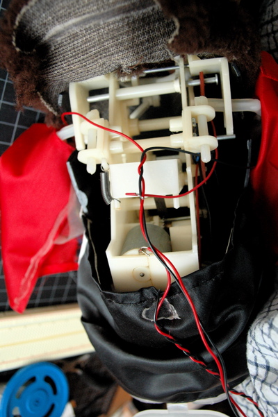Photo of a toy monkey. The back has been removed to reveal the inner gear mechanism that plays the cymbals. At the center of a mechanism is a DC motor. Wires have been attached to it to run the motor from an H-bridge.