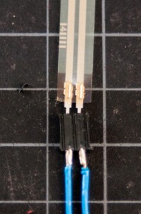 Photo of the metal ends of a force-sensing resistor mounted in breakaway socket headers. The socket headers, separated by 0.1 inches (2.5mm) have wires soldered to their metal ends. This way, the soldering happens to the socket pins. The socket headers are cheaper than the sensor, so the cost of damaging them by soldering is less than soldering the sensors. 