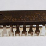 This component is a small rectangle with eight pins on each of the two long sides. There is a small round depression on the upper left corner of the rectangle, and a semicirular dip in the middle of the top.