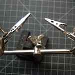Photo of helping hands tool. The tool has a metal base witha swivel joint at the top. The swivel joint holds a metal bar approximately 6 inches (15cm) long. Each end of the bar has another swivel joint with a metal clip mounted on it.