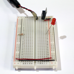 Photo of a solderless breadboard with a 7805 voltage regulator mounted on it, as in the images above. The regulator is connected to a DC power plug, and the ground and voltage output of the regulator is connected to the voltage and ground bus rows on the right side of the breadboard. The ground and voltage bus rows on the right are connected to the ground and voltage bus rows on the left with wires at the bottom of the board. A 220-ohm resistor is connected to the voltage bus on the left. Its other end is connected to row twelve in the center area. An LED is connected to a hole in row twelve on the right side. The other side of the LED is connected the ground bus on the right side.