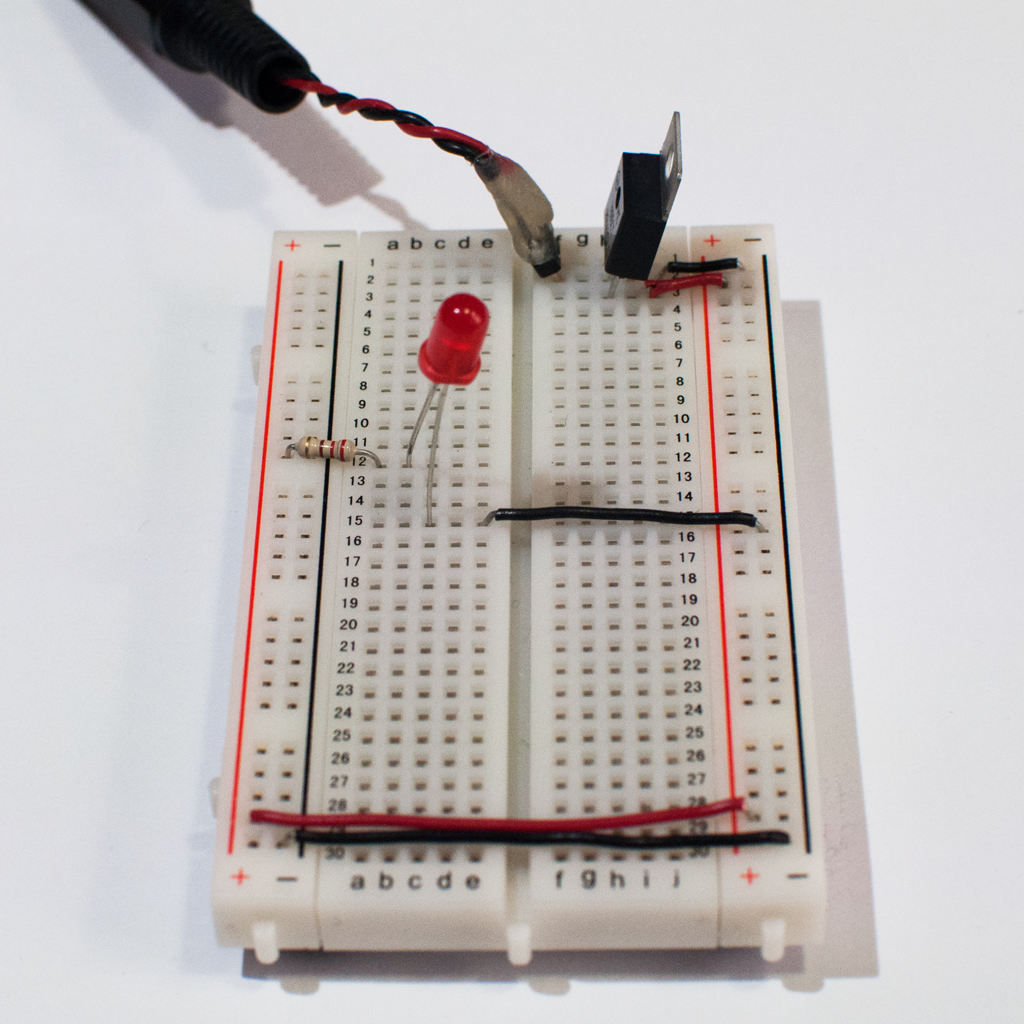 Photo of a solderless breadboard with a 7805 voltage regulator mounted on it, as in the images above. The regulator is connected to a DC power plug, and the ground and voltage output of the regulator is connected to the voltage and ground bus rows on the right side of the breadboard. The ground and voltage bus rows on the right are connected to the ground and voltage bus rows on the left with wires at the bottom of the board. A 220-ohm resistor is connected to the voltage bus on the left. Its other end is connected to row twelve in the center area. An LED is connected to another hole in row twelve. The other side of the LED is connected to a hole in row fourteen. Another hole in row fourteen connects to the ground bus on the right side of the board.