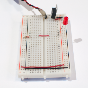 Photo of a solderless breadboard with a 7805 voltage regulator mounted on it, as in the images above. The regulator is connected to a DC power plug, and the ground and voltage output of the regulator is connected to the voltage and ground bus rows on the right side of the breadboard. The ground and voltage bus rows on the right are connected to the ground and voltage bus rows on the left with wires at the bottom of the board. A 220-ohm resistor is connected to the voltage bus on the left. Its other end is connected to row thirteen in the center area. A red wire connects row thirteen on the left with row thirteen on the right. An LED is connected to a hole in row thirteen on the right side. The other side of the LED is connected the ground bus on the right side.