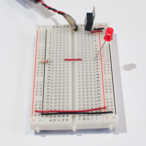 Photo of a solderless breadboard with a 7805 voltage regulator mounted on it, as in the images above. The regulator is connected to a DC power plug, and the ground and voltage output of the regulator is connected to the voltage and ground bus rows on the right side of the breadboard. The ground and voltage bus rows on the right are connected to the ground and voltage bus rows on the left with wires at the bottom of the board. A 220-ohm resistor is connected to the voltage bus on the left. Its other end is connected to row thirteen in the center area. A red wire connects row thirteen on the left with row thirteen on the right. An LED is connected to a hole in row thirteen on the right side. The other side of the LED is connected the ground bus on the right side. The LED is lit up.