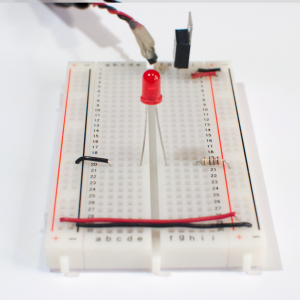 Photo of a solderless breadboard with a 7805 voltage regulator mounted on it, as in the images above. The regulator is connected to a DC power plug, and the ground and voltage output of the regulator is connected to the voltage and ground bus rows on the right side of the breadboard. The ground and voltage bus rows on the right are connected to the ground and voltage bus rows on the left with wires at the bottom of the board. A 220-ohm resistor is connected to the voltage bus on the right. Its other end is connected to row twenty in the on the right side of the center area. An LED is connected to a hole in row twenty on the right side as well. The other side of the LED is connected row twenty on the left side. A black wire connects row twenty on the left with the ground bus on the left side.