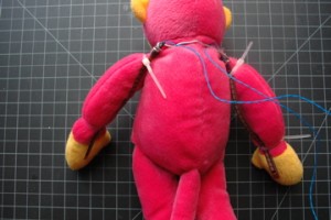 Photo of a stuffed pink monkey with flex sensors attached to its arms. The flex sensors are attached with tie-wraps at the shoulder and wrist of the monkey. The wired ends of the sensors are behind the monkey's neck. The wires lead away from the monkey's back discreetly.