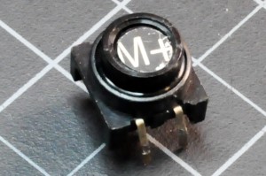 Photo of a small round button, approximately 0.5 in (2-3cm) in diameter. The button has two legs on either side. Pushing the button closes the connection between the two legs on each side.
