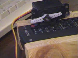 Photo of a remote control mounted in a wooden cradle. A servomotor mounted on the side of the cradle is positioned such that when it moves, its horn presses down on the power button of the remote control.