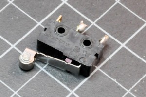 Photo of a rocker switch. A rectangular component approx. 1 in (2-3cm) wide by 0.25 in (0.5cm) thick. A lever is mounted on one side, and three metal contacts on the other. Under the lever is a small plastic protrusion that is pushed down when the lever is pressed.