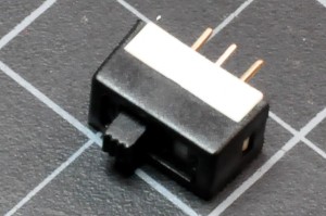 Photo of a slide switch. A component approx. 1.5cm wide by 0.5cm thick. There are three metal legs on one side, and a plastic handle at the top that can slide from one side to the other. Sliding the handle switches the center leg's connection from one side leg to the other.