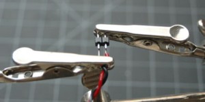 Photo of two header pins held in a helping hands tool. The headers are touching two wires, one red, one black. The wires and the header pins are not soldered together. 