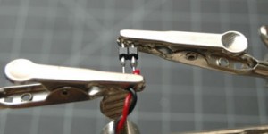 Photo of two header pins held in a helping hands tool. The headers are touching two wires, one red, one black. The wires and the header pins are soldered together. 