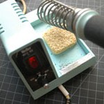 Photo of a soldering iron. The base of the iron is approximately 5 inches (13cm) on a side. The left side is reaised and has a power switch and a temperature knob on the front. The right side has a metal coil, and the iron itself is inserted in the coil. The iron is approximately 8 inches (20cm) long, metal with a plastic handle. The handle has a thick wire coming out of the back that attaches to the base below the temperature knob.