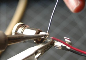 Photo of a wire hooked into the metal tab of a power connector. Both are held by a pair of helping hands clips. A soldering iron is touched to the joint between the wire and the tab. A piece of solder is touched to the joint, but is not touching the iron directly.