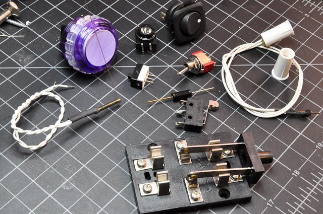 An assortment of switches and pushbuttons. This image includes an arcade-style pushbutton; an old typewriter key; a round toggle switch; a small; rocker switch; a whisker switch, which is a thin wire attached to a spring wrapped around a metal post; a magnet switch, and a magnet that closes the switch when you bring it close; a tilt switch; a two-way knife switch. 
