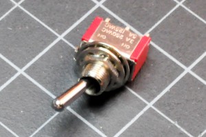 Photo of a toggle switch. A component approx. 1.5cm wide by 0.5cm thick. There are three metal legs on one side, and a metal lever at the top center that can move from one side to the other. Moving the lever switches the center leg's connection from one side leg to the other.