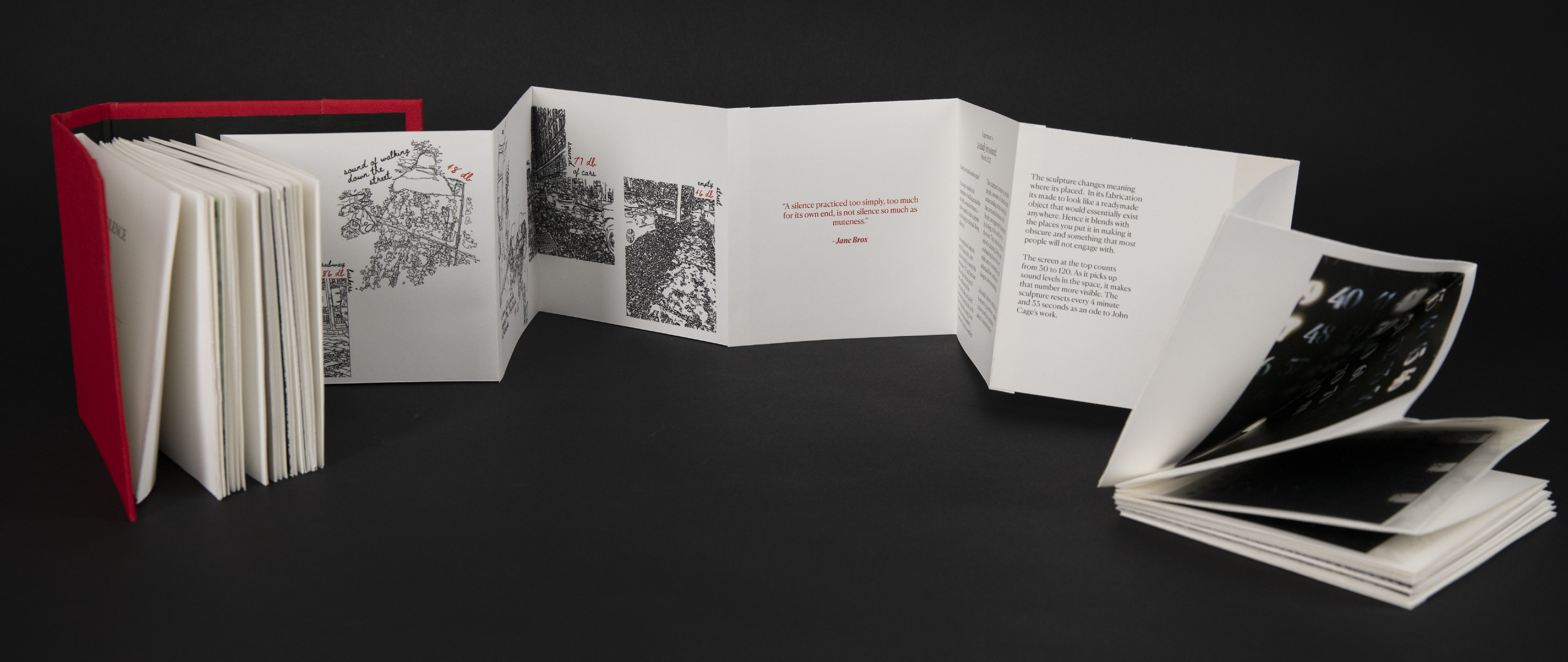 Red book placed at an angle with pages facing right corner of the screen. Pages are attached as an accordion, part of the accordion has been pulled out of the book and a part of the middle of the book is placed in a zigzag pattern, a one-third end of the book is in a stack on the right edge of the frame.
