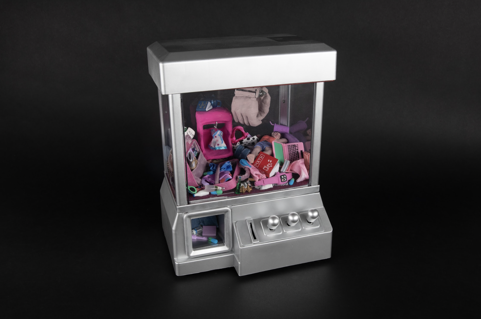 A claw machine game filled with doll size clutter. A sculpture of a person in bed lies beneath the clutter.