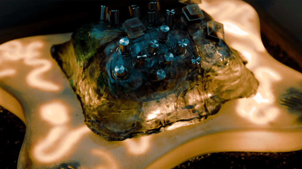 homemade hardware synth on a bed of soil and rocks. glowing mossy greens and blues, blobous forms, and warm squiggling light.