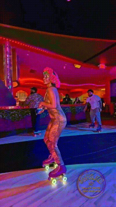 Rollergirl is smiling, standing up in a brown galatatic speckled body suit on a pair of purple rollerskates in a  roller rink