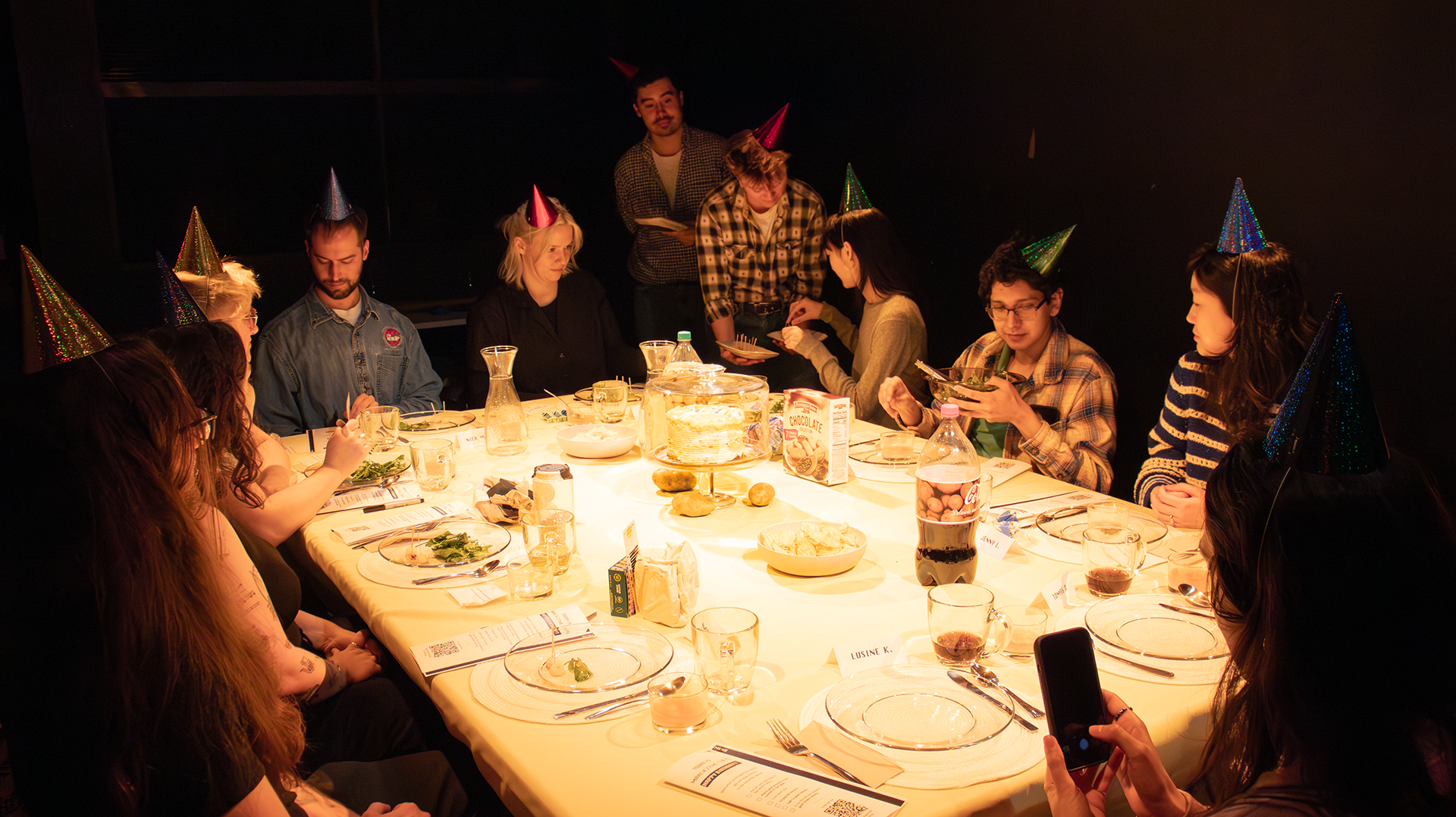 Ten guests with party hats are seated around a large table. Two facilitators are passing around food.