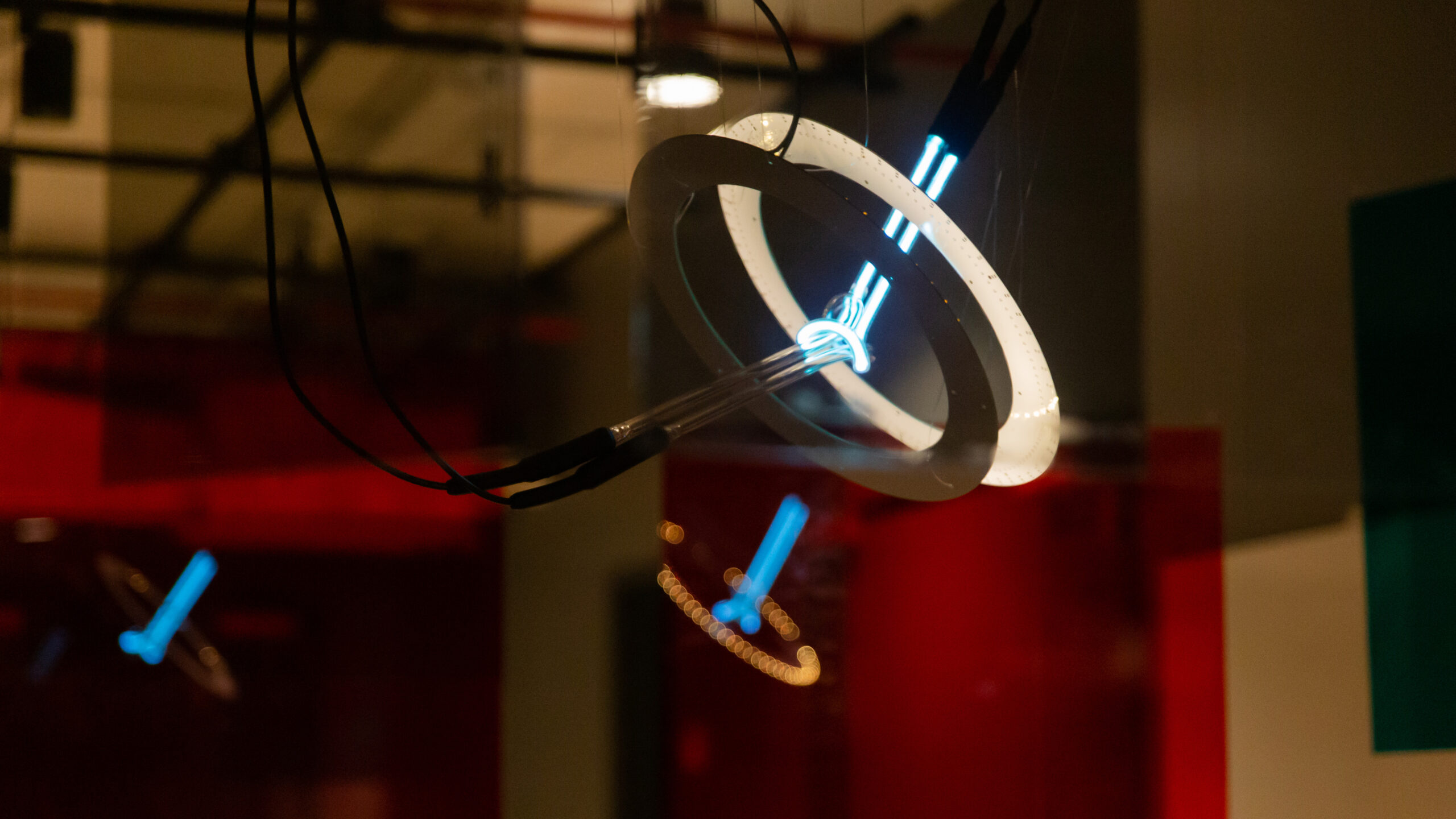 The light sculpture with only one neon piece lights up in blue, surrounded by two white, ring-shaped printed circuit boards. One circuit board is lit up in white by the LEDs on the other circuit board. The entire sculpture is seen through a layer of polarized filter, while its reflections can be seen on the red color panels in the background.