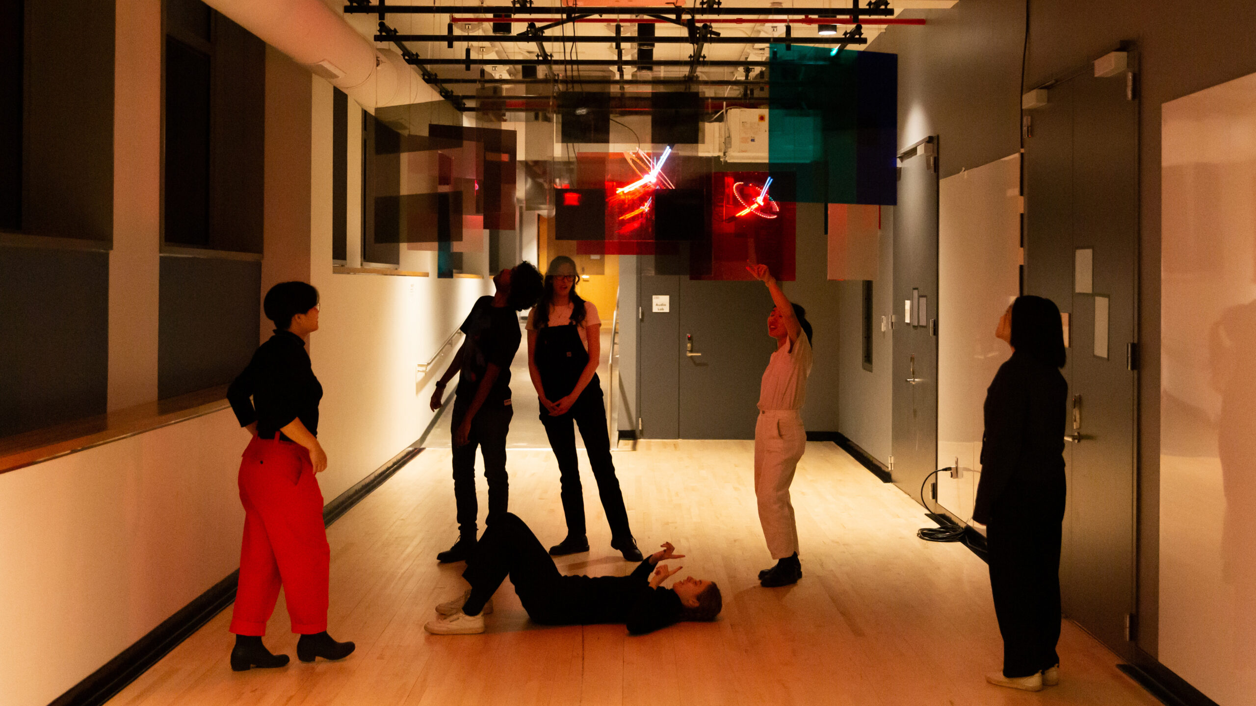 Six of my friends are interacting with the installation. From left to right: Leia is standing outside the perimeter of the acrylic panels, gazing at the light sculpture; Suraj is underneath the mirror panel in the back and turning his head to look at the reflection; Zoe is leaning towards Suraj while looking at Martha, who is lying on the floor, looking up at the light sculpture while gesturing a camera frame; Julia is pointing at a panel and seems to be conversing with Martha, Zoe, and Leia; I-Jon is standing outside the perimeter to the right, gazing at the light sculpture.