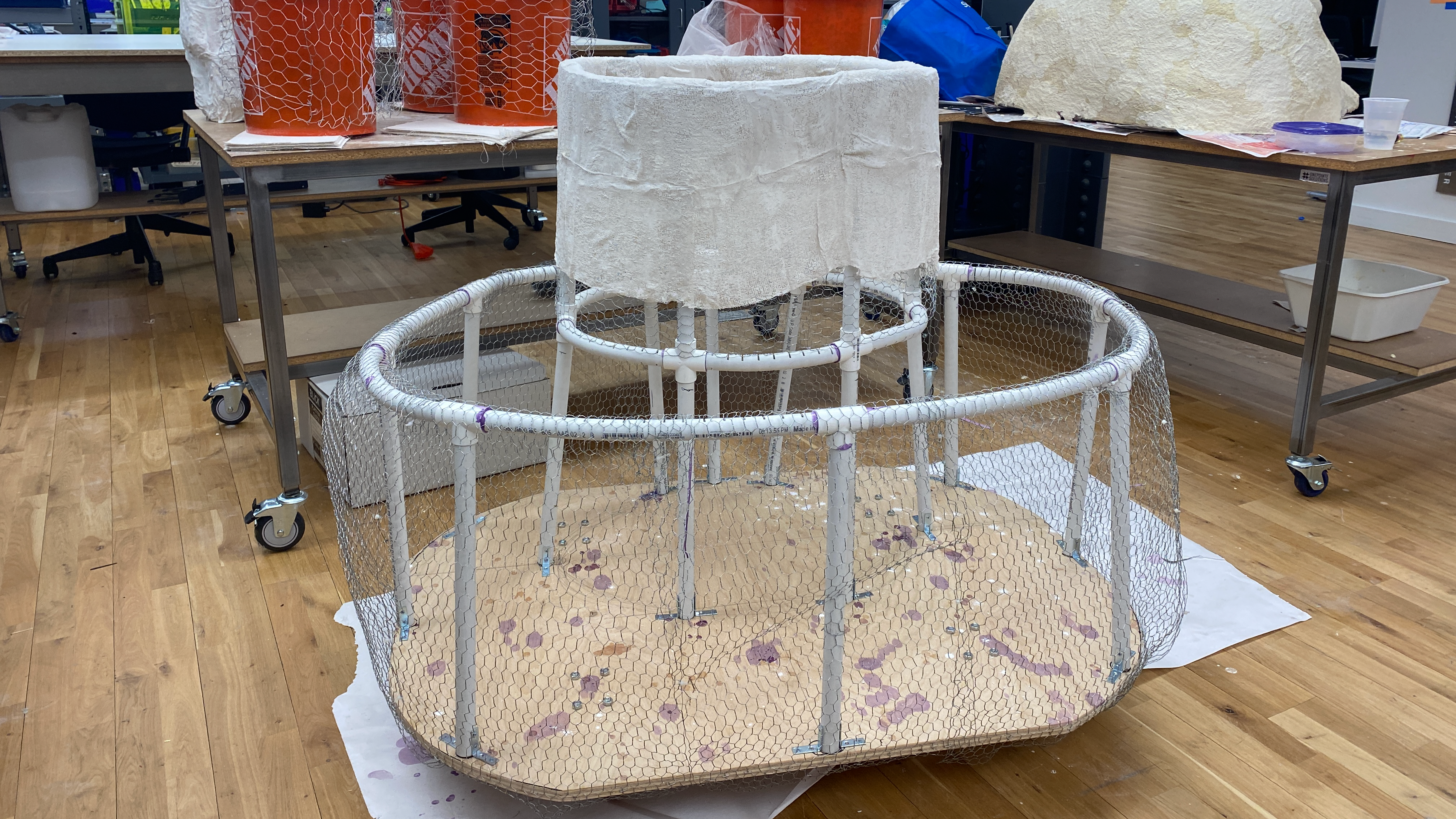 an oval frame of bent PVC pipes covered in a wire mesh and attached to a wood board sits in the middle of a workspace. A center smaller tower inside the larger frame is partially covered with plaster cloth