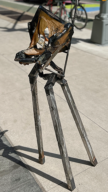 A three legged steel sculpture on a day lit skywalk. The upper part of the sculpture has a brownish/yellowish leathery skin adhered to it.
