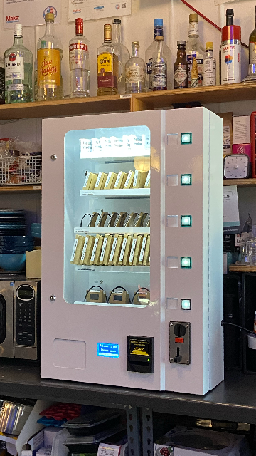 A white full vending machine displaying on a kitchen shelf. There are bottles of alcohol and random kitchen items in the background.