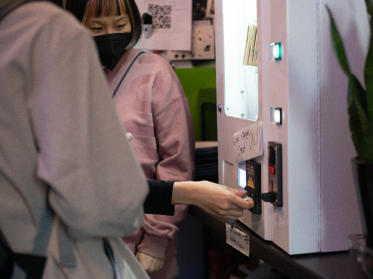 People interacting the Ultimate Vending Machine by inserting cash to the machine. Someone in the back are speculating the interaction.